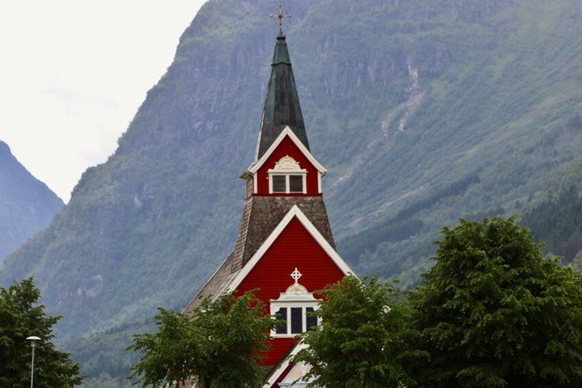 A red church with a steep, pointed steeple, surrounded by lush greenery and mountains. 3