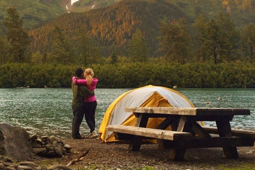 A couple embracing by a lake with a tent and picnic table. 1