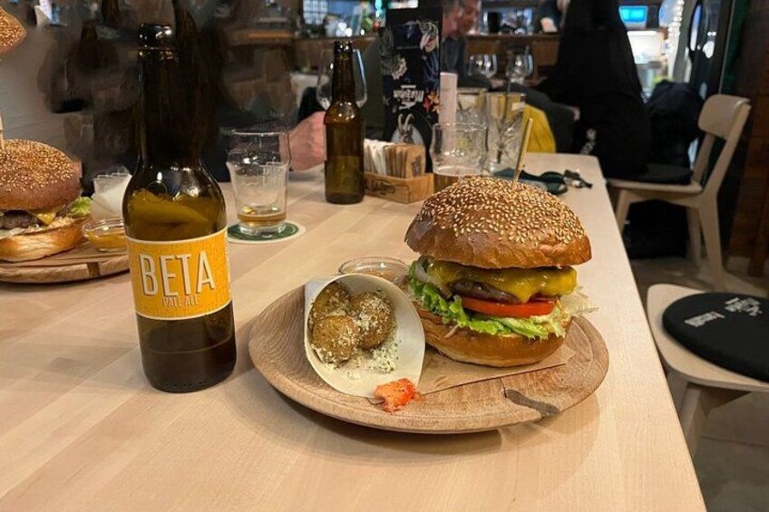 A burger with a sesame seed bun, a bottle of Beta Pale Ale, and a small plate with a food item on a wooden table. 2