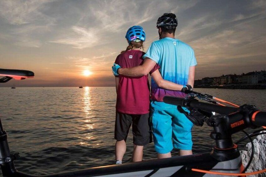 Two people standing on a boat, a sunset in the background, and a city skyline across the water. 1
