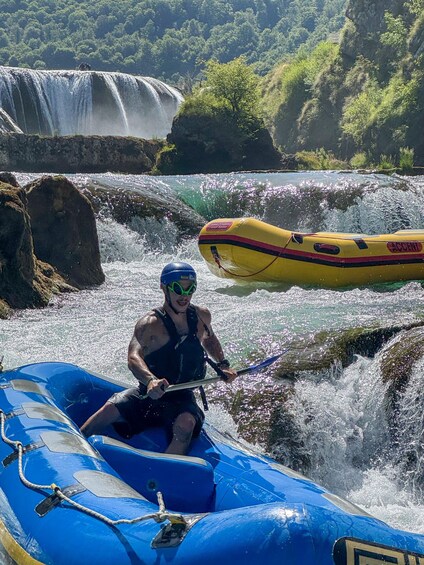A person in a blue raft navigating rapids, with a yellow raft and a waterfall in the background. 4