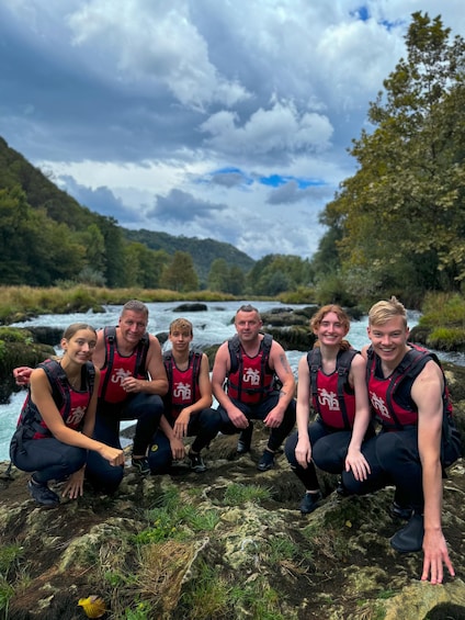 A group of people in red life jackets posing on a rocky outcrop by a flowing river, surrounded by greenery and hills. 3