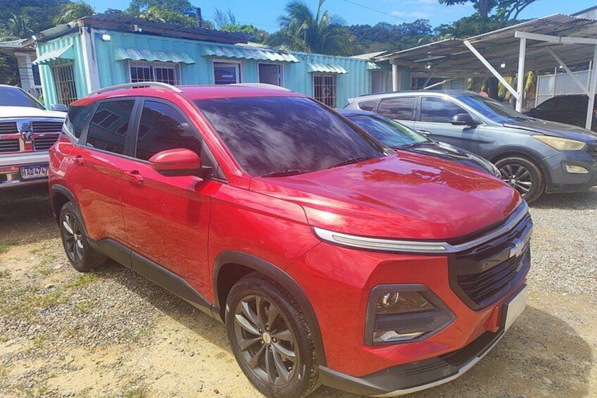 A red SUV parked on a gravel surface with buildings in the background. 5