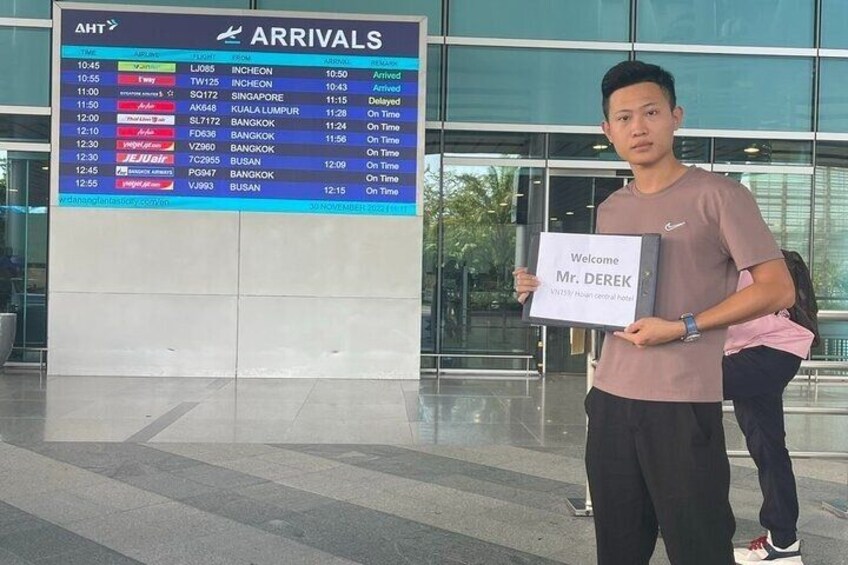 A man holding a welcome sign in an airport arrivals area with a digital flight information board in the background. 2