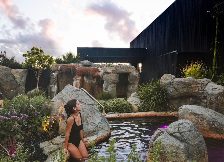 A woman in a black swimsuit sitting on a rock by a pond with a waterfall and a modern building in the background. 1