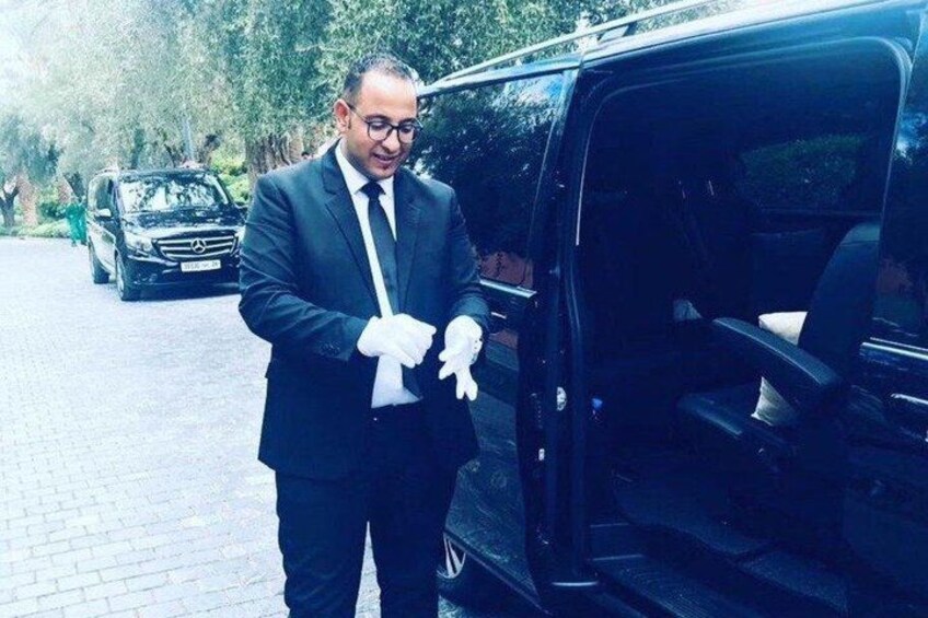 A man in a suit standing next to a black van with a car in the background. 4