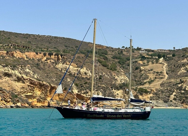 Ein Segelboot namens 'Sua Maesta' mit der Aufschrift 'Rosa dei Venti' auf ruhiger See, im Hintergrund felsige Hügel und ein klarer Himmel. 2