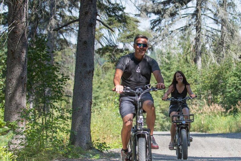 Two people riding bicycles on a paved path surrounded by trees. 1