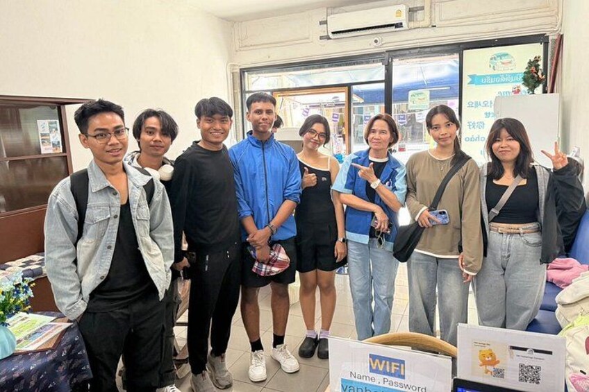 A group of people standing indoors, some with backpacks, in a room with a table, a box labeled 'WIFI', and a window with a view of the outside. 3