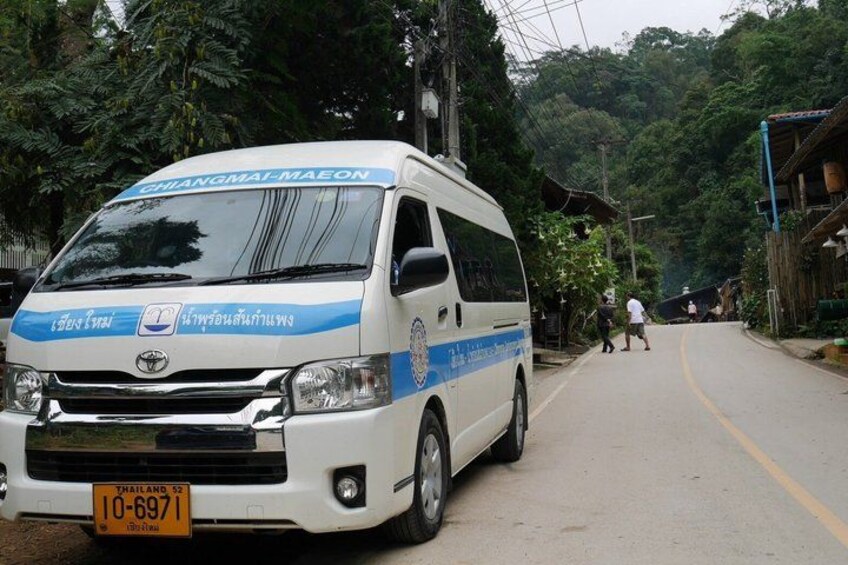 A white and blue bus with Thai text on the front, parked on a road with people walking in the background. 1