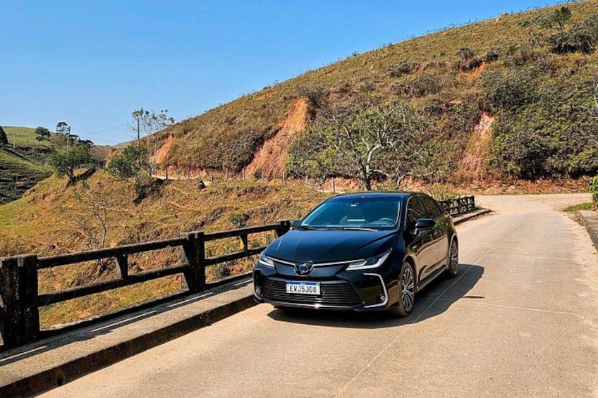 A black car parked on a road with a wooden guardrail on the left, a hilly landscape in the background, and a clear sky. 2