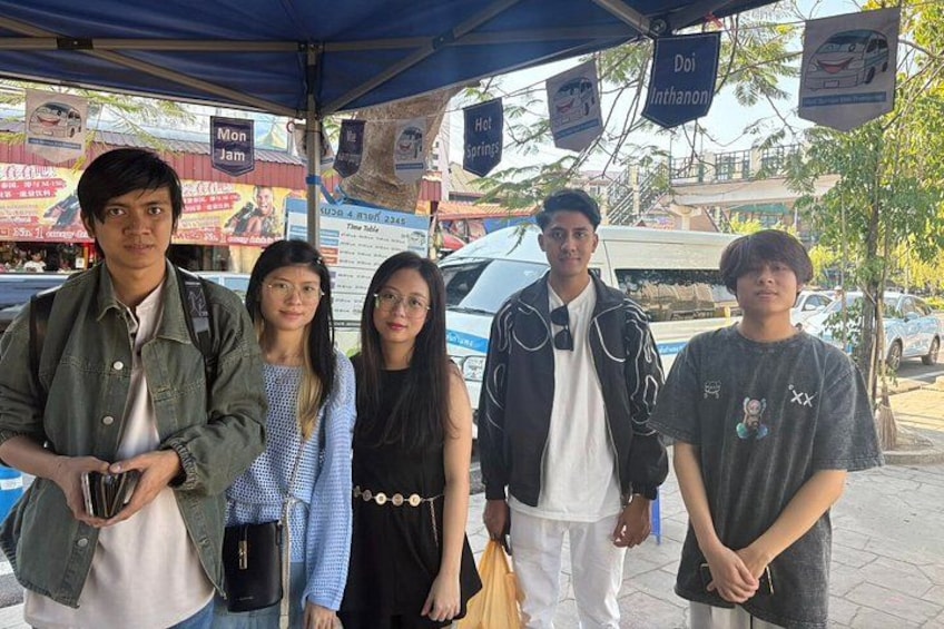A group of people standing under a blue canopy with signs advertising a restaurant and a car parked in the background. 3