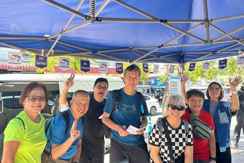 A group of people under a blue canopy with signs for 'Hot Springs' and 'Deli Intha'. 2