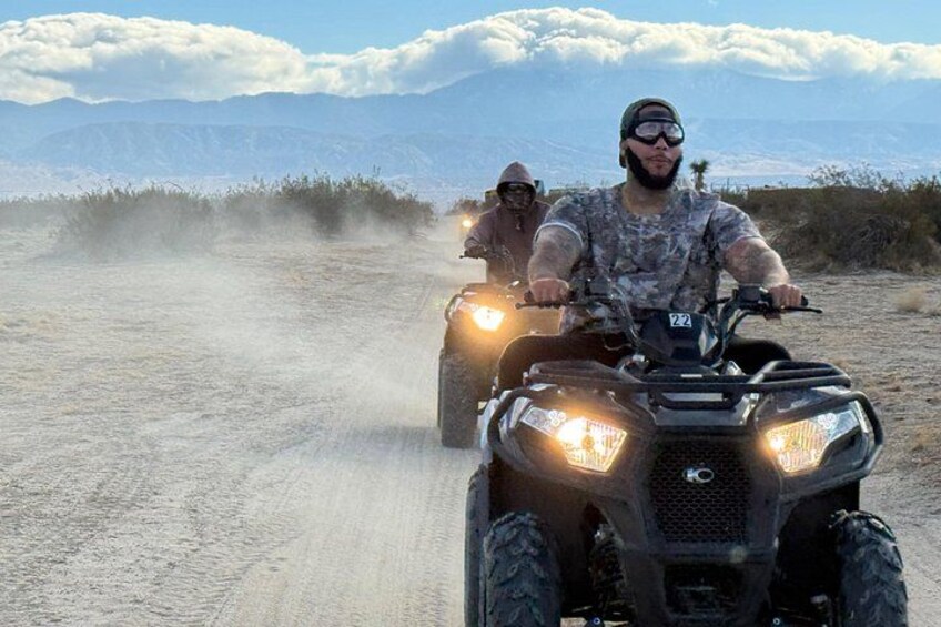 Two people riding ATVs on a dusty road with mountains in the background. 4