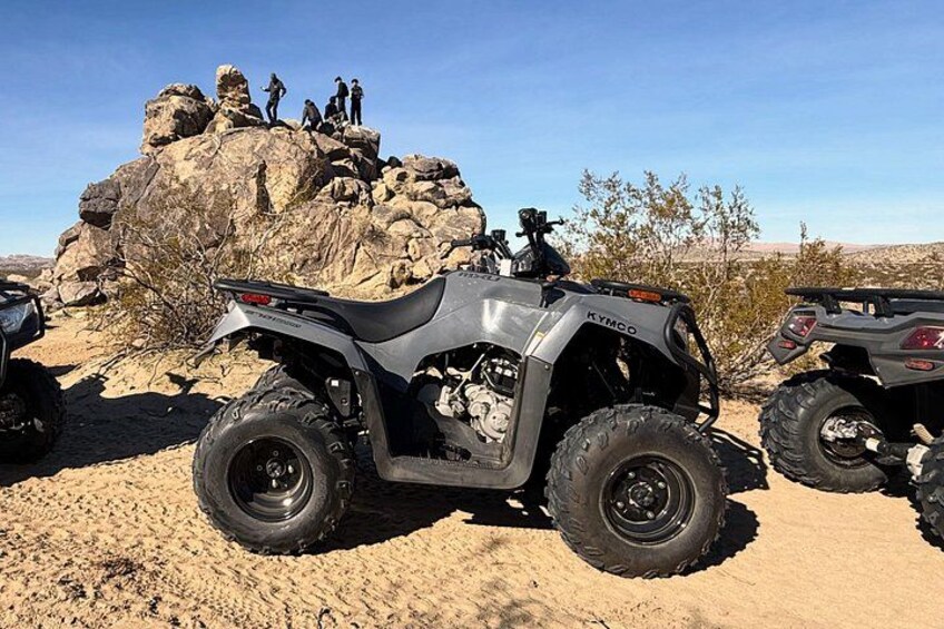 A quad bike parked on a sandy terrain with rocky formations and sparse vegetation in the background. 1