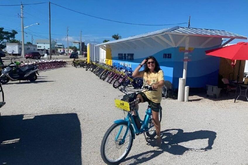 A person on a blue bicycle with a yellow basket, parked on a gravel road with a row of parked motorcycles in the background. 4