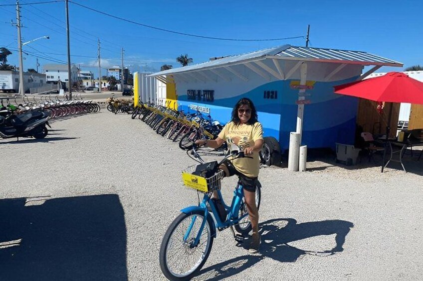 A person standing next to a blue bicycle with a yellow basket, parked on a gravel road with a row of parked bicycles and a small building with a blue and white color scheme in the background. 3