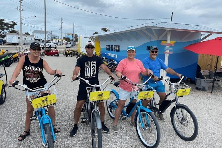 Three people on bicycles with baskets, parked in a lot with a blue building in the background. 1