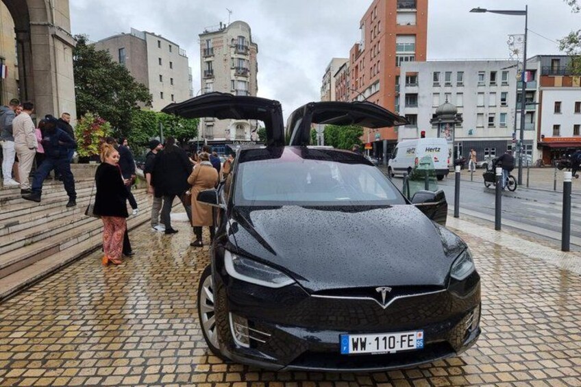 A black car parked on a cobblestone street with its trunk open, surrounded by pedestrians and buildings. 5