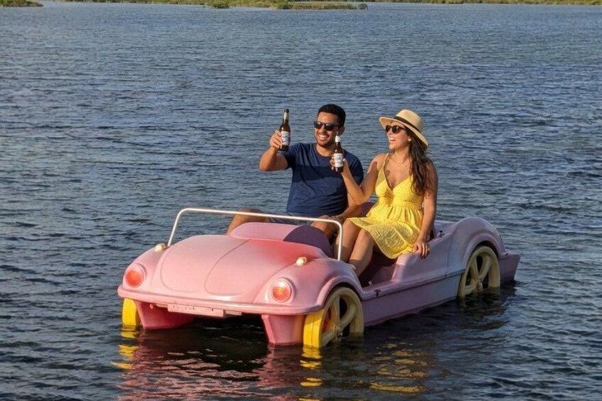 Two people on a pink boat with a car-like design on a body of water. 1