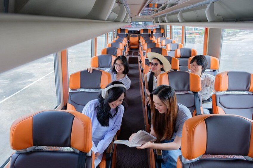 A bus interior with orange and gray seats, passengers seated, and a window showing a road outside. 1