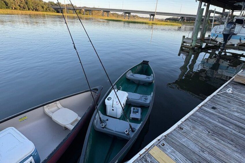A small green boat docked at a wooden pier with calm water and a bridge in the background. 2