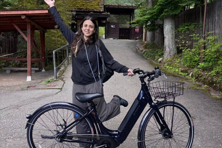 A person standing next to a stationary electric bicycle with a basket, in a paved area with a wooden structure and greenery in the background. 5