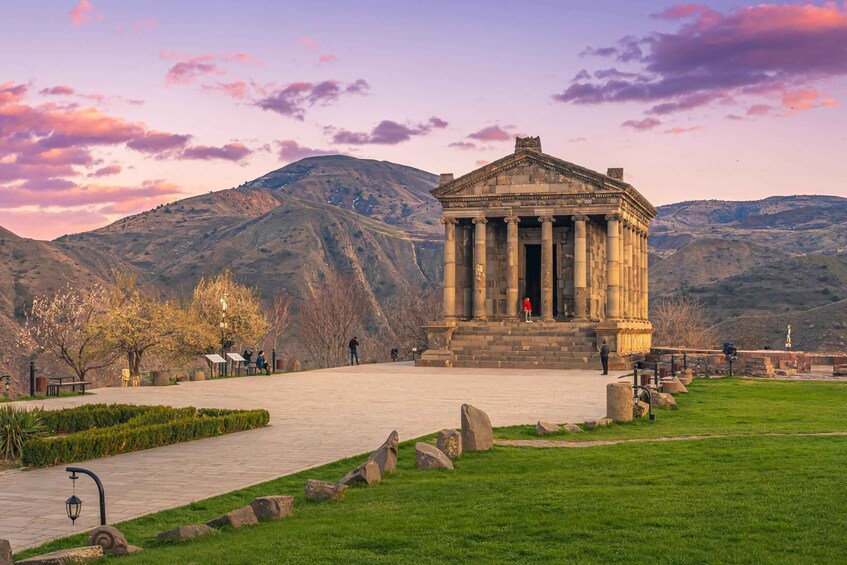 Un edificio neoclásico con columnas, rodeado por un parque con bancos y un sendero, con montañas al fondo durante el atardecer. 1