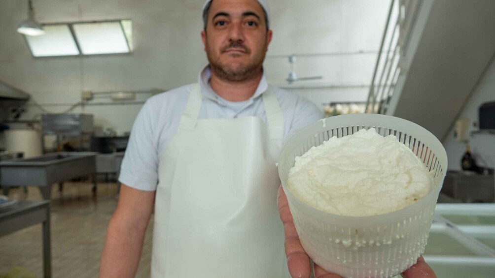 Una persona con delantal blanco sostiene un colador lleno de una sustancia blanca, probablemente harina, en una cocina con electrodomésticos de acero inoxidable y una ventana. 3