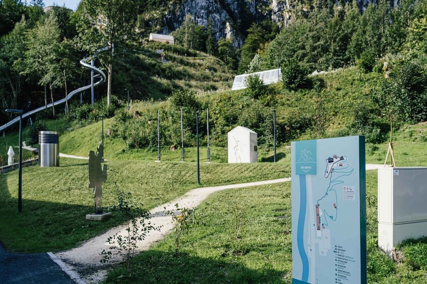 Un sentier de randonnée, une sculpture métallique, un panneau indicateur avec une carte, et un bâtiment situé dans une zone vallonnée. 3