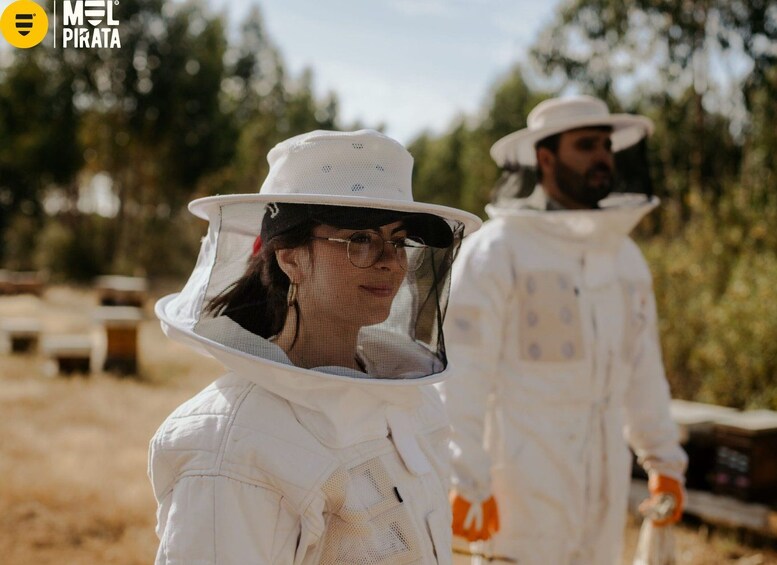Two individuals in protective white suits and hats, likely engaged in an outdoor activity, with trees and a wooden structure in the background. 3