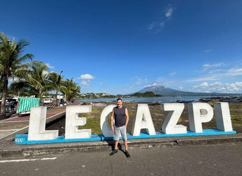 Un hombre de pie frente a un cartel con la palabra 'LEGAZPI' y vista a una playa con barcos y una montaña al fondo. 2