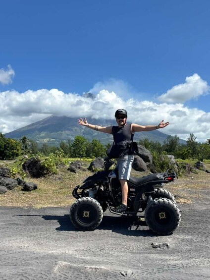 Una persona parada en un cuatrimoto con los brazos extendidos, una montaña con nubes al fondo y un cielo despejado. 4