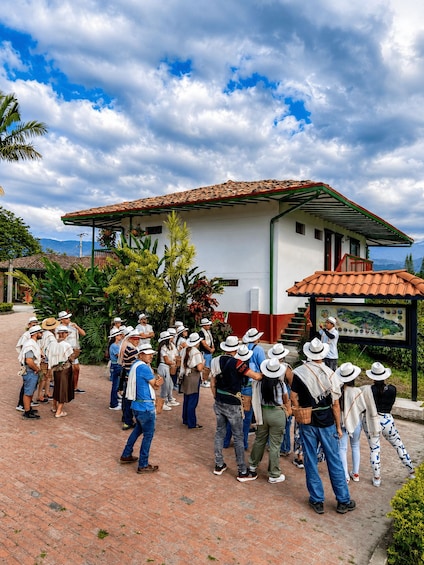 A group of people gathered around a building with a red-tiled roof, standing on a brick-paved area with greenery and a mountainous backdrop. 1
