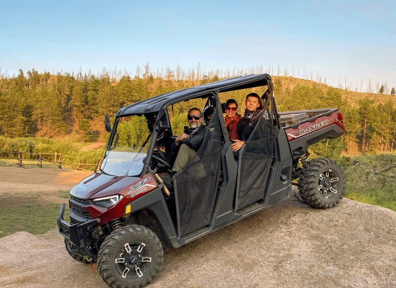 A black off-road vehicle with four passengers, parked on a concrete surface with a forested hillside in the background. 1