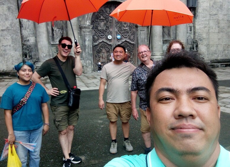Eine Gruppe von Personen steht vor einem Gebäude mit aufwendigen Schnitzereien und hält Regenschirme. 5