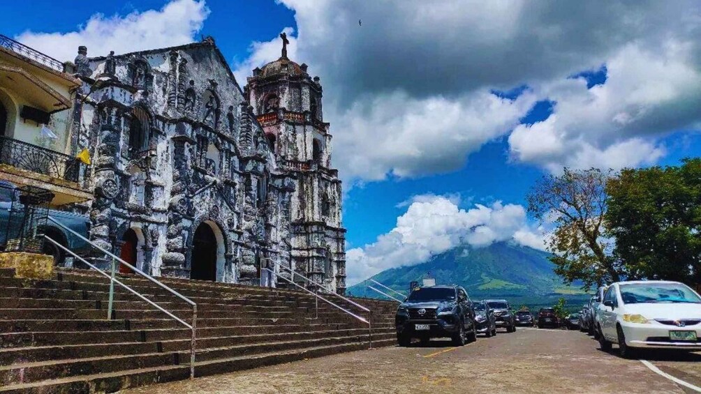A historic church with a bell tower, steps leading up to it, parked cars, and a mountain in the background. 2