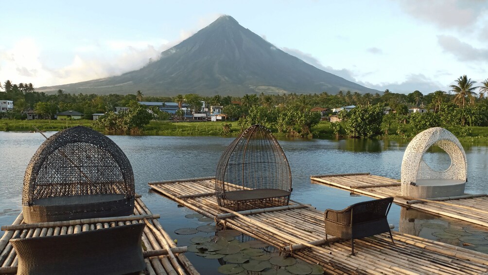 A bamboo dock with woven chairs and a white structure by a calm lake, with a mountain in the background. 4