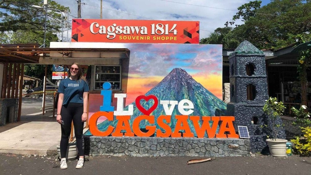 A person standing in front of a sign that reads 'Cagsawa 1814 Souvenir Shoppe' with a large 'I Love Cagsawa' sign in the background. 5