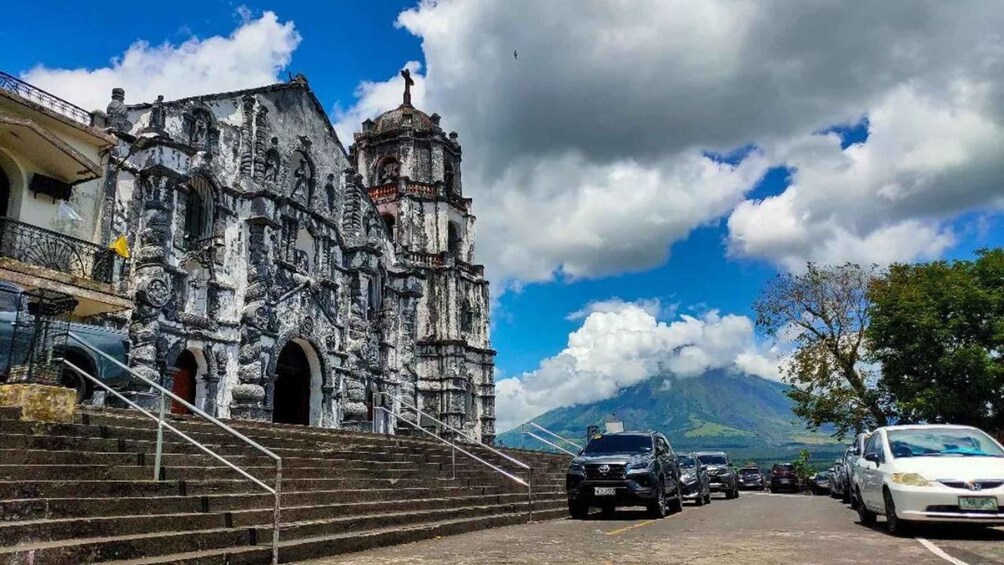 A historic church with a bell tower, a staircase leading up to it, parked cars, and a mountain in the background. 3