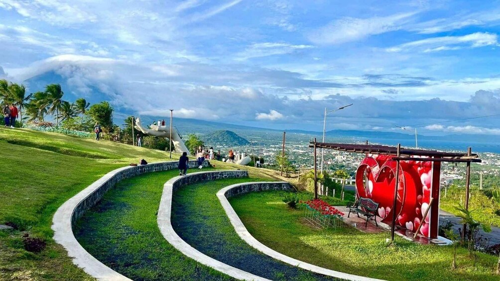 A park with winding pathways, a red heart-shaped structure, and a view of a city and mountains in the distance. 2