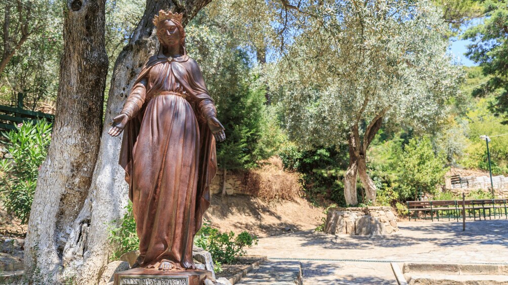 Una estatua de bronce de una mujer con vestido largo, de pie sobre un pedestal con una placa, rodeada de árboles y un muro de piedra en un parque. 3