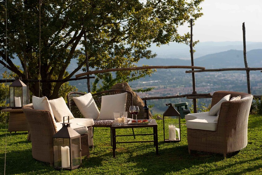 An outdoor seating area with wicker furniture, a table with a candle, wine glasses, and a bottle, set against a backdrop of a large tree and a view of distant mountains. 3