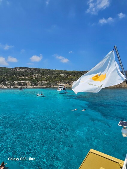 Mar azul y transparente con barcos, una bandera y un paisaje montañoso al fondo. 4