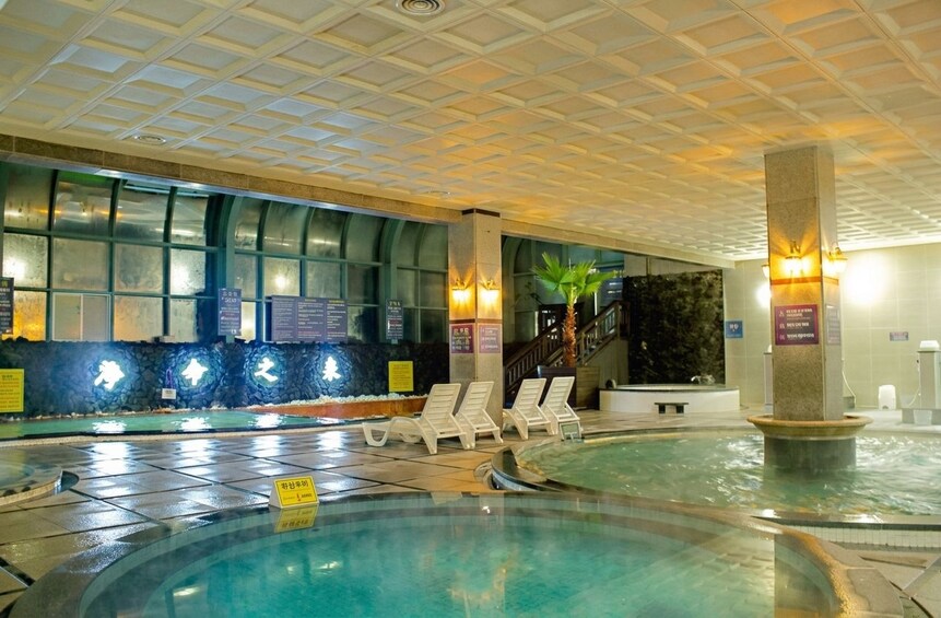 Ein Innenpool mit Liegestühlen, eine Steinwand mit Wasserspielen und eine Treppe, die zu einer erhöhten Ebene führt. 5