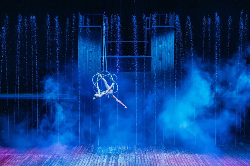 A performer in a hoop suspended above a stage with water jets and mist. 1