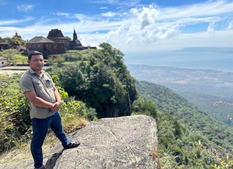 A man standing on a rock with a view of a temple and a valley. 1