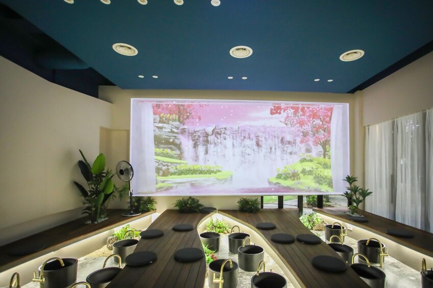 A modern interior with a large projection screen displaying a waterfall scene, wooden tables with black stools, and potted plants. 3