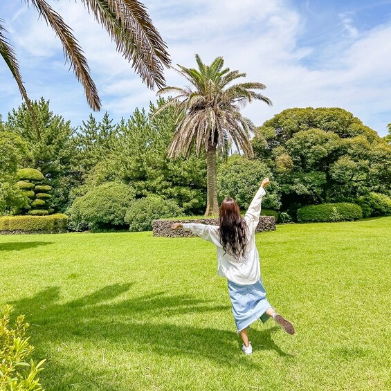 A person in a white shirt and blue skirt dancing in a lush green park with palm trees and neatly trimmed bushes. 1