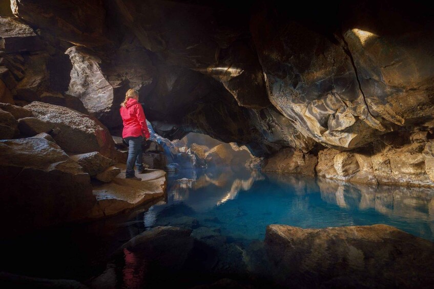 Une personne vêtue d’une veste rouge, debout sur un rocher, près d’une piscine aux eaux bleues et limpides, dans une grotte rocheuse. 3